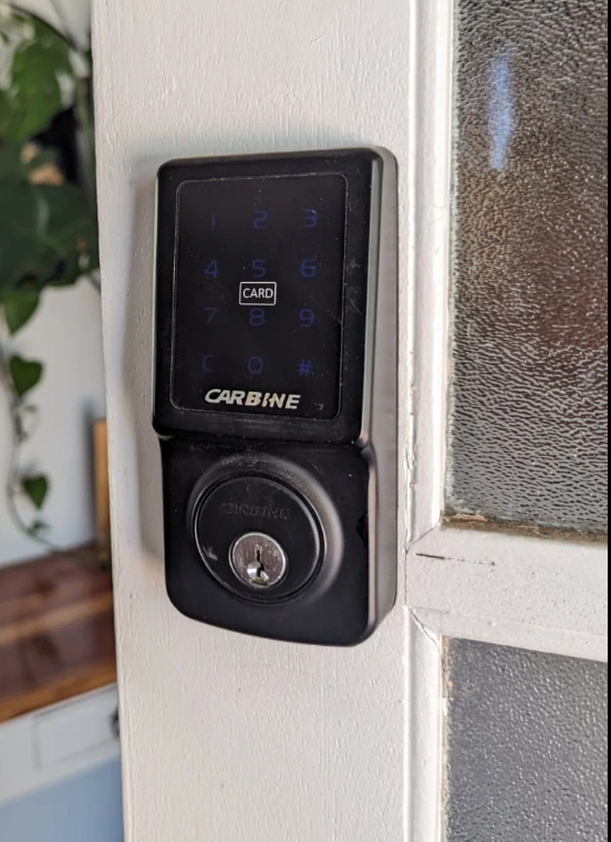 A locksmith’s gloved hand holding a new silver deadbolt lock against a wooden residential door, demonstrating an on-site lock replacement.